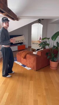 Caixa de cartão com rótulo "zalando" segurada por uma pessoa em roupa casual. O ambiente apresenta um sofá de veludo côtelê laranja e uma grande planta em vaso. Pavimento de madeira.