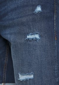 Läheltä otettu kuva sinisistä farkuista, joissa on kolme kulutettua ja purjehtimista reikää reisi-alueella ja näkyvää oranssia ompelua.