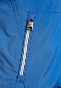 Tessuto della giacca a conchiglia morbida blu con una cerniera bianca visibile e un cursore argentato, caratterizzata da cuciture a zigzag e una texture liscia.