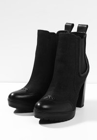 Botas negras hasta el tobillo con un upper de tela texturizada y una puntera de cuero suave, con un detalle decorativo de tachuelas y un tacón cuadrado.