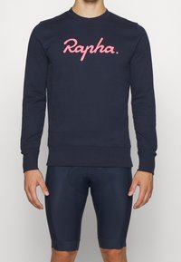 Felpa navy con logo "Rapha." rosa, collo a giro e polsini a costine. Indossata con shorts da ciclismo navy aderenti, che mostrano un design slanciato.