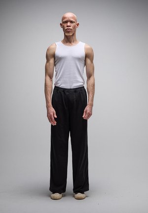 Man met albinisme die een wit mouwloos shirt, zwarte wijde broek en beige schoenen draagt, staand tegen een effen grijze achtergrond.