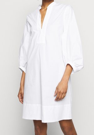 Vrouw die een loszittende witte jurk draagt met lange pofmouwen en een V-hals, staand tegen een effen lichte achtergrond.