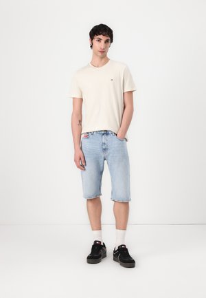 T-shirt en coton beige avec un petit logo, associé à un short en denim bleu clair et des baskets noires avec des chaussettes blanches. Tenue simple et décontractée.