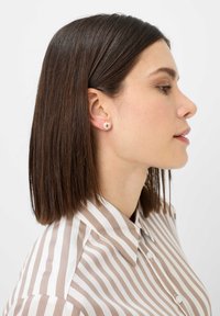 Orecchini a bottone in oro rosa con un design circolare caratterizzato da un motivo a onda. Il modello indossa una camicia a righe beige e bianca, con i capelli lisci e lunghi fino alle spalle.
