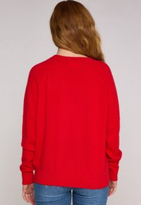 Maglione rosso lavorato a maglia con una vestibilità rilassata, maniche raglan e orlo a coste. La texture sembra morbida e accogliente, adatta per un abbigliamento casual.