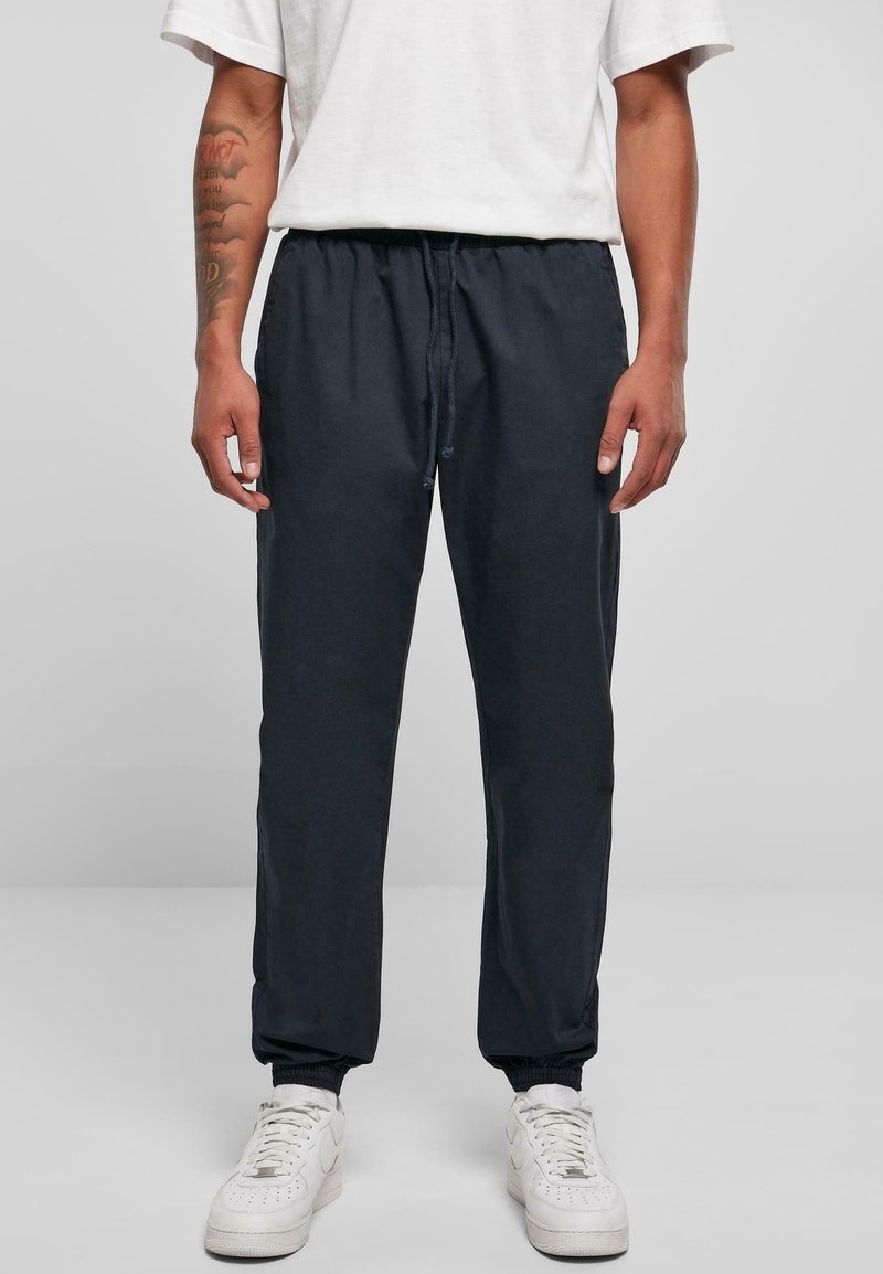 Personne portant un pantalon de jogging noir avec des poignets élastiques, des baskets blanches et un T-shirt blanc uni, debout devant un fond uni.