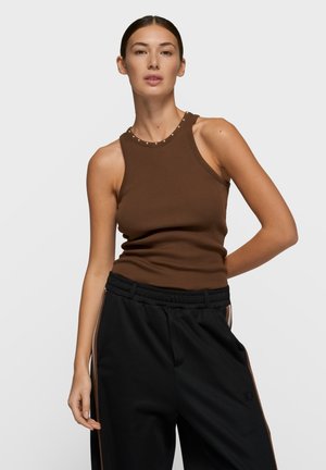 Brown ribbed tank top with a round neckline accented by metallic studs, paired with black loose-fit trousers featuring a side stripe.