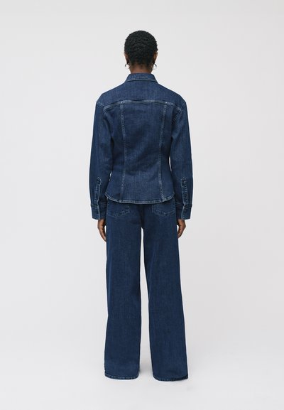 Giacca in denim blu scuro con silhouette aderente, caratterizzata da maniche lunghe, polsini con bottoni e colletto classico, abbinata a pantaloni in denim a gamba larga.
