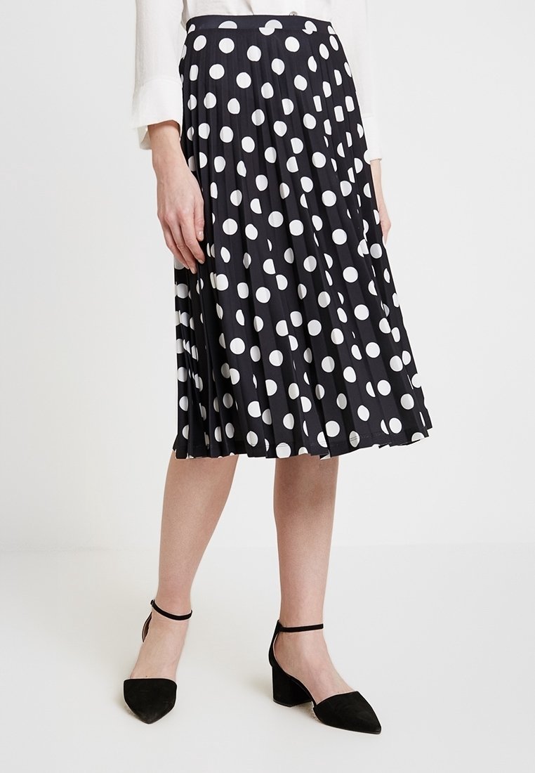 Femme portant une jupe midi plissée noire à gros pois blancs et des talons carrés noirs avec bride à la cheville.
