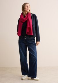 Marinejakke, sort top, brede blå jeans, hvide sneakers med sorte detaljer, og et bølget pink tørklæde draperet om halsen.