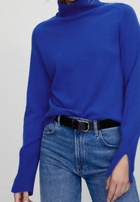 Jasnoniebieski sweter typu golf z miękkiego materiału, z długimi rękawami z otworami na kciuki, w połączeniu z jasnoniebieskimi dżinsami i czarnym paskiem.