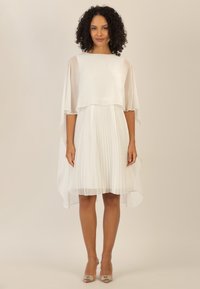 Abito bianco con gonna a pieghe, maniche a cappuccio semitrasparenti e scollatura rotonda. Accentuato da décolleté beige.