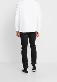 Camisa blanca de manga larga con corte recto, con puños abotonados. Llevada con jeans ajustados negros y zapatillas de lona negras, mostrando un dobladillo enrollado.