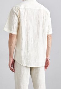 Lyhythihainen paita, joka on valmistettu teksturoidusta kermaisen värisestä kankaasta. Siinä on kaulus, rento leikkaus ja kaareva helma, yhdistettynä vaaleankirjaviin housuihin.