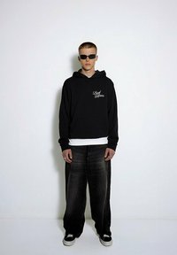 Czarna bluza z kapturem z napisem "Lead Moves", nałożona na białą koszulkę; w zestawie z luźnymi, ciemnymi dżinsami i czarnymi sneakerami. Model nosi okulary przeciwsłoneczne.