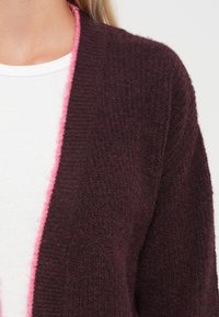Gilet en tricot dans un bordeaux foncé avec des bordures roses. Texture douce, design à front ouvert. Une chemise blanche en dessous est partiellement visible.