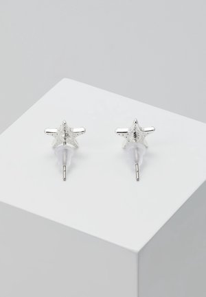 Silberne sternförmige Ohrstecker mit klaren Kunststoffrückseiten, präsentiert auf einer weißen, schrägen Oberfläche.