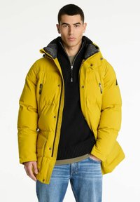 Gul puffjakke med grå hætte, lynlås foran og flere lommer, lagret over en sort strikket sweater og synlige jeans.