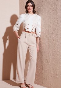 White cropped shirt with embroidered floral patterns; beige high-waisted wide-leg trousers; sleek sandals; textured wall background.