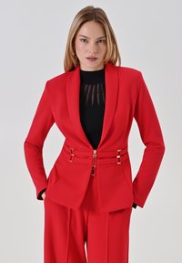 Rode blazer met een getailleerde pasvorm, voorzien van gouden hardware accenten en een cinched taille. Gecombineerd met een zwart geribbeld topje, voor een slanke uitstraling.