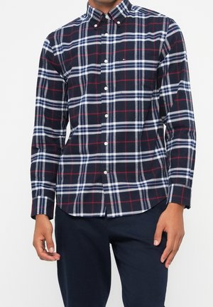 Muž oblečený v námořnicky modré, bílé a červené kostkované košili s dlouhými rukávy a námořnicky modré kalhoty, stojící před bílým pozadím.