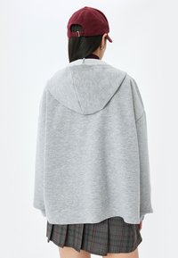 Lys grå hættetrøje med en løs pasform og struktureret stof. Bæres over en ternet nederdel. En burgunder cap fuldender outfittet.