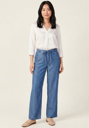 Femme debout portant un chemisier blanc à col en V, un jean bleu ample à cordon, et des mocassins beiges, sur un fond clair uni.