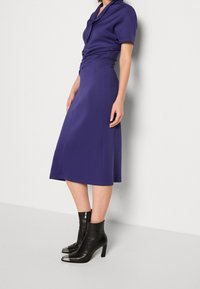Fialové midi šaty s krátkým rukávem, s úzkým pasem a rozšířenou sukní. Nosené s černými kotníkovými botami s kovovými špičkami a zipem.