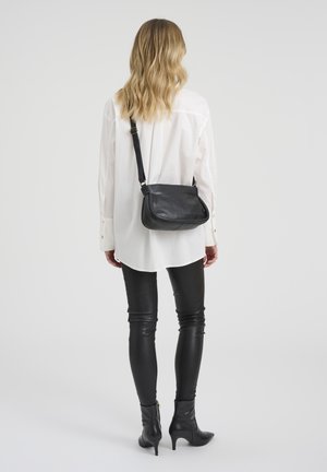 Žena s valovitom plavom kosom, u bijeloj majici s dugim rukavima, crnim kožnim hlačama, crnim čizmama s potpeticom i crnom torbom preko tijela, okrenuta leđima.