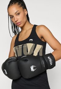 Mustat nyrkkeilyhanskat, joissa valkoinen logo. Käyttäjällä on yllään musta hihaton paita, jossa on kultaisia kirjaimia ja raidallinen kuvio, ja paita on vartalonmyötäinen.