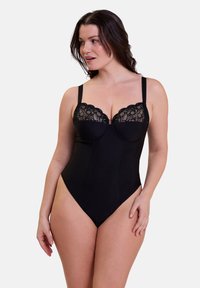 Femme portant un body noir avec des bonnets en dentelle, se tenant contre un fond clair, regardant sur le côté avec une main sur la cuisse.