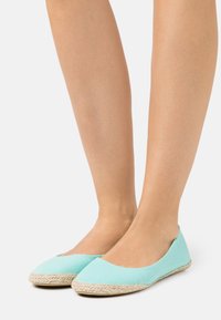 Sapatilhas de tela azul claro com bico redondo, apresentando uma sola de juta trançada. O design é minimalista, sem fechos ou adornos visíveis.