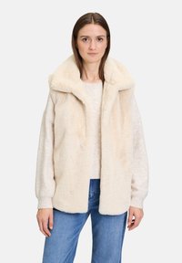 Beige Faux-Fur-Weste mit großem Kragen, langen Ärmeln und lockerer Passform, kombiniert mit einem leichten Pullover und blauen Jeans.