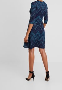 Polvenmittainen mekko tummansinisenä, jossa on turkoosi siksak-kuvio, pitkät hihat ja levenevä helma. Yhdistetty mustien korkokenkien kanssa.