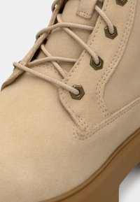 Beige semsket støvel med høy topp, med sekskantede metalløye, flate lisser og gummisåle. Glatt tekstur og rene sømdetaljer.