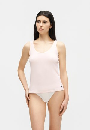 Γυναίκα με μακριά σκούρα μαλλιά, που φορά ροζ αμάνικη μπλούζα και λευκό εσώρουχο, στέκεται μπροστά σε μονόχρωμο φόντο.