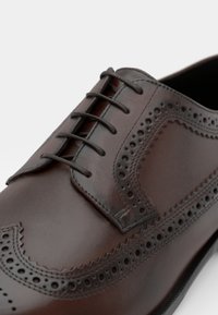 Zapato de vestir de cuero marrón con diseño de brogue, superficie lisa, punta afilada y seis ojales para los cordones, con detalles decorativos perforados.
