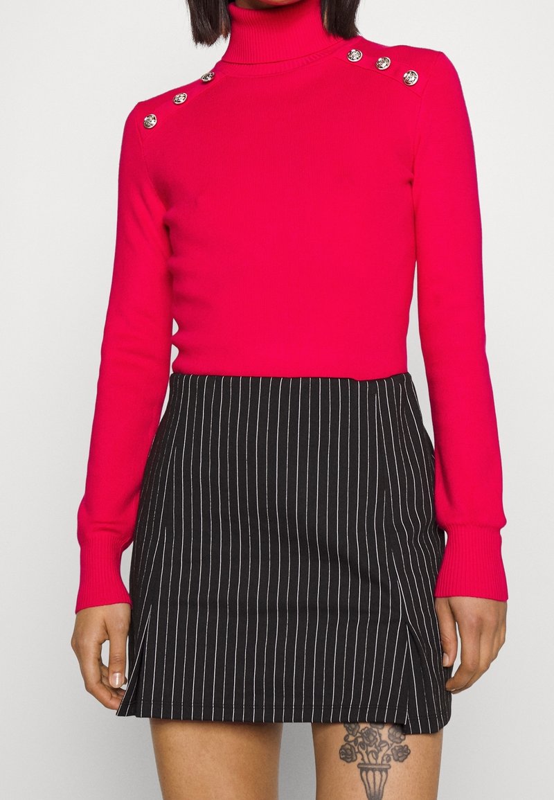 Enganliegender rosa Rollkragenpullover mit dekorativen silbernen Knöpfen an den Schultern, kombiniert mit einem schwarzen Minirock mit Nadelstreifen. Strukturierter Strickstoff.