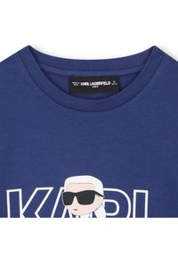 Temno modra bombažna majica s kratkimi rokavi, na kateri je risani lik z sončnimi očali in napis "KARL" v beli barvi. Ovratnik z rebrastim zaključkom.