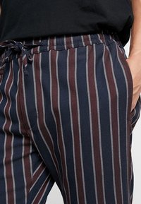Pantalons rayés avec un schéma de couleurs bleu marine et bordeaux, ornés de rayures verticales blanches. La taille dispose d'un cordon de serrage ajustable pour un meilleur ajustement.