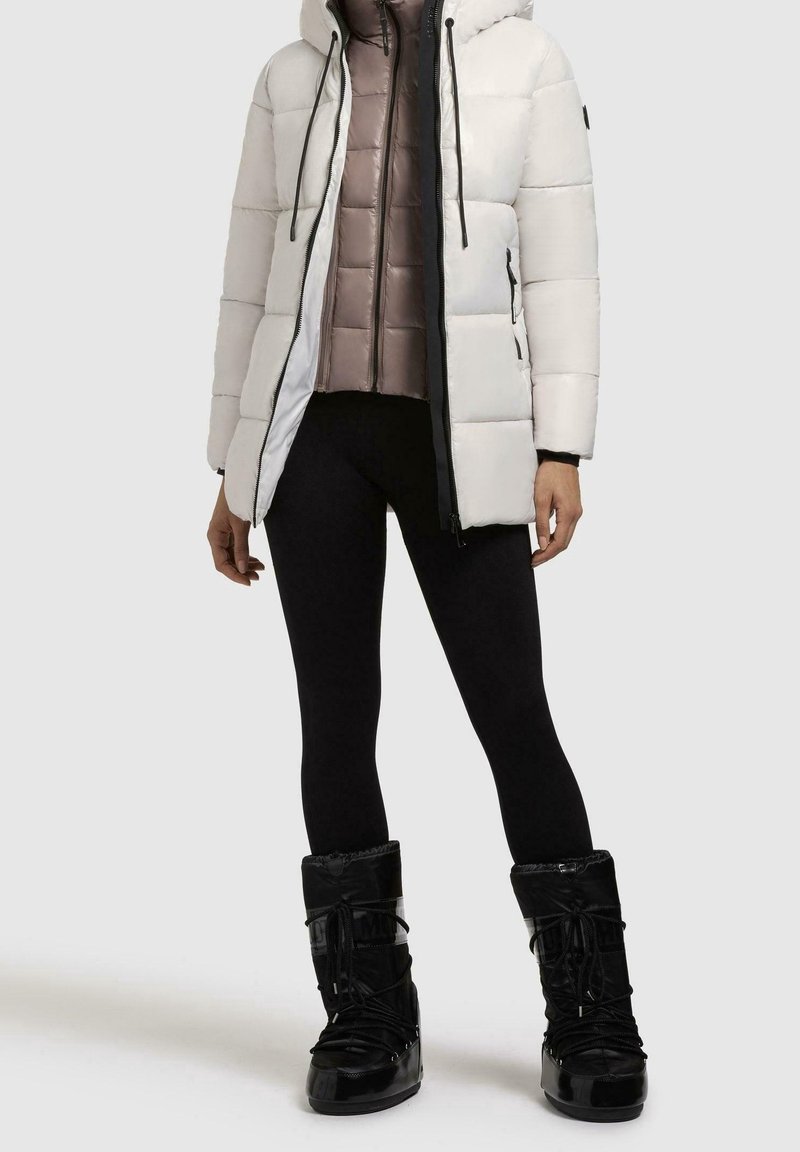 Chaqueta blanca tipo puff con capucha y detalles negros, superpuesta a un chaleco acolchado rosa claro. Leggings negros y botas negras brillantes completan el atuendo.