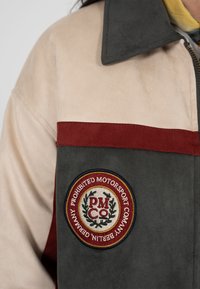 Section de veste avec tissu supérieur beige, tissu inférieur gris foncé, bande horizontale rouge et écusson circulaire "Prohibited Motorsport Company".