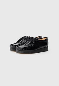 Black patent leather shoes with a rounded toe, decorative stitching, and a low textured wedge sole. Features matching laces for closure.