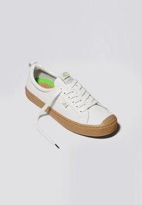 Sneaker bianco in tela con suola in gomma color caramello, accenti verdi all'interno e un logo sulla linguetta. Presenta lacci piatti e un design casual.