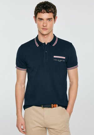 Młody mężczyzna w granatowej koszulce polo z czerwono-białym paskowanym kołnierzem i beżowych spodniach, stojący z jedną ręką w kieszeni na jednolitym tle.