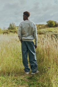 Ung person i grå sweatshirt og blå jeans, der står på en græsklædt marksti og vender væk under en skyet himmel.