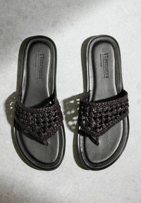 Paire de sandales noires sans lacets avec des brides tissées et un entre-doigts, reposant sur une surface texturée gris clair.