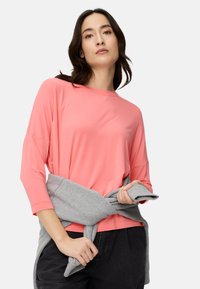 Koralfarvet langærmet top med afslappet pasform, rund halsudskæring og sænkede skuldre; gråt striktrøje bundet om taljen; båret med mørke bukser.