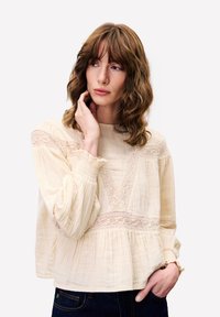 Frau mit welligem braunem Haar, die eine cremefarbene Spitzenbluse und dunkle Jeans trägt, sich ins Gesicht fasst und leicht zur Seite schaut.